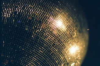 A close-up of a disco ball reflecting purple and black lights, casting playful sparkles across a dark dance floor.