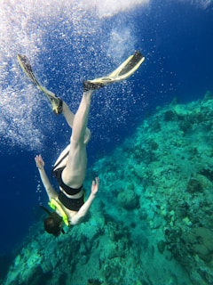 A person wearing a snorkel and fins is diving underwater near a coral reef. The clear blue water and sunlight create a vibrant and serene underwater scene.