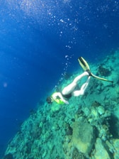 A person is snorkeling underwater, surrounded by clear blue water and a rocky seabed. The individual is wearing a snorkel, mask, and bright yellow flippers, and is positioned head-down, exploring the underwater environment. Bubbles rise around them, and the sunlight filters through the water, creating a serene atmosphere.