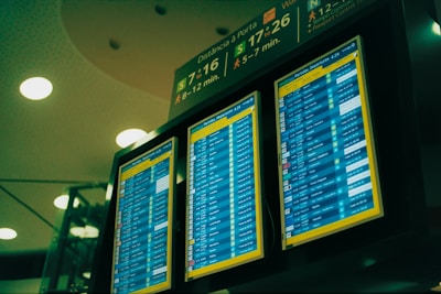 A set of three digital display screens showcases departure information at an airport. The screens are brightly lit with text and numerical data, indicating flight details such as times and gate numbers. Overhead signage provides directions to passengers, with estimated walking times to various gates.