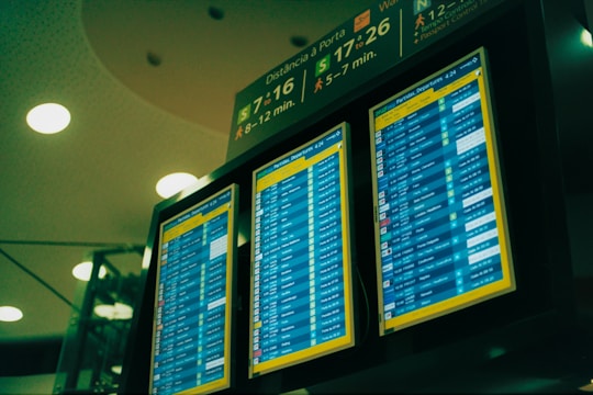A set of three digital display screens showcases departure information at an airport. The screens are brightly lit with text and numerical data, indicating flight details such as times and gate numbers. Overhead signage provides directions to passengers, with estimated walking times to various gates.