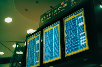 A set of three digital display screens showcases departure information at an airport. The screens are brightly lit with text and numerical data, indicating flight details such as times and gate numbers. Overhead signage provides directions to passengers, with estimated walking times to various gates.