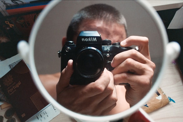 A person is holding a classic Nikon camera and taking a picture of themselves in a round mirror. Various items are scattered on the table including a passport, a boarding pass, and other documents.