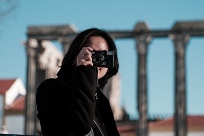 A person is holding a camera up to their face, capturing a moment. The background features the blurred image of ancient ruins with columns, and the sky is clear.