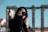 A person is holding a camera up to their face, capturing a moment. The background features the blurred image of ancient ruins with columns, and the sky is clear.