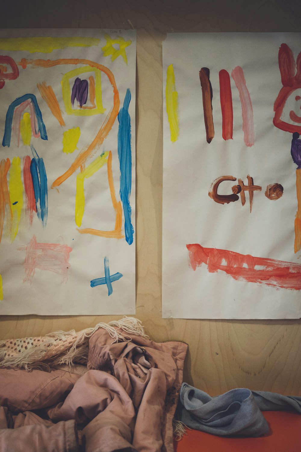 two children's paintings on a wall next to a pile of clothes