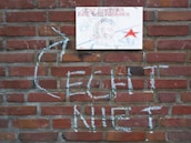 A brick wall serves as the backdrop with a piece of paper or small poster attached to it. The poster features the phrase 'LUCKY BUSTERS ARE VICTORIOUS' in a bold, decorative font above a drawing of a character with a helmet, possibly intended to be humorous or satirical. A prominent red star is also part of the design. Below the poster, words and symbols are scrawled on the wall in white chalk.