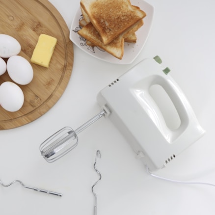 A kitchen scene featuring a white electric hand mixer surrounded by a wooden board with three eggs and a rectangular piece of butter. A pile of toasted bread is placed on a plate nearby, and there are mixer attachments scattered on the white countertop.