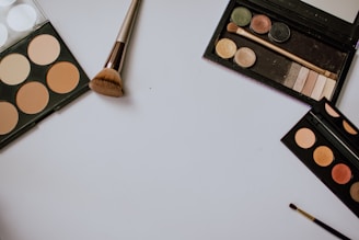 A neatly arranged setup of makeup items featuring several palettes with different shades, including a contour palette, an eyeshadow palette, and a small brush. The items are laid out on a white surface, with a larger makeup brush positioned centrally.