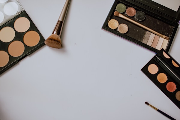 A neatly arranged setup of makeup items featuring several palettes with different shades, including a contour palette, an eyeshadow palette, and a small brush. The items are laid out on a white surface, with a larger makeup brush positioned centrally.
