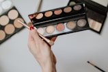 A hand with red nail polish is holding an eyeshadow palette with a brush. The palette contains various shades of shimmery colors. Other makeup palettes are visible in the background, possibly containing contour or foundation shades.