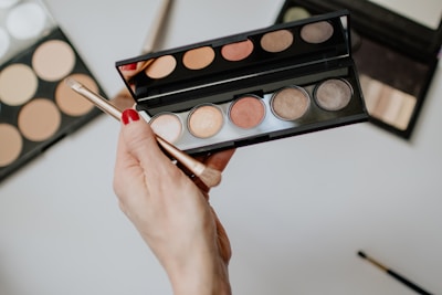 A hand with red nail polish is holding an eyeshadow palette with a brush. The palette contains various shades of shimmery colors. Other makeup palettes are visible in the background, possibly containing contour or foundation shades.
