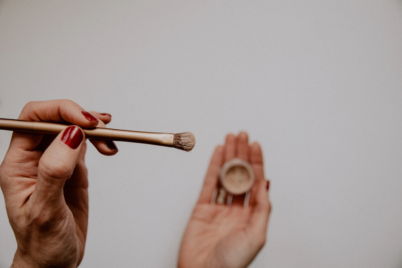 One hand holding a makeup brush above a small pot of loose pigment held in the other hand