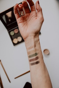 An extended arm with makeup swatches on the inner forearm. The swatches are in earthy tones, including shades of brown and green. A makeup palette and a brush are visible in the background.