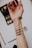 An extended arm with makeup swatches on the inner forearm. The swatches are in earthy tones, including shades of brown and green. A makeup palette and a brush are visible in the background.