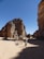 A caravan of camels led by a person traverses a sandy desert landscape, flanked by towering rocky cliffs under a clear blue sky.