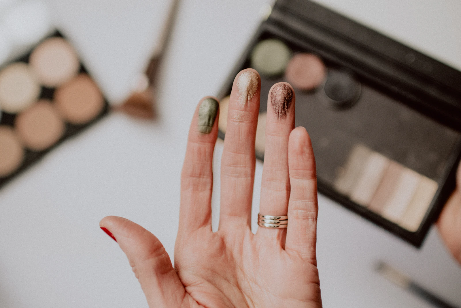 A hand with eyeshadow swatched on the fingertips, an open palette behind it