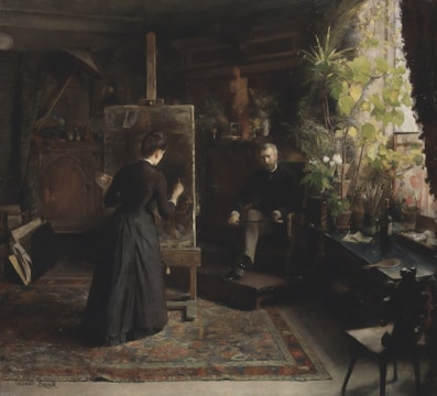A dark, richly detailed art studio with an artist painting at an easel. The room is filled with natural light from a large window, highlighting the lush greenery of potted plants. A seated man observes the artist's work, surrounded by various objects including brushes, a table, and a patterned rug.