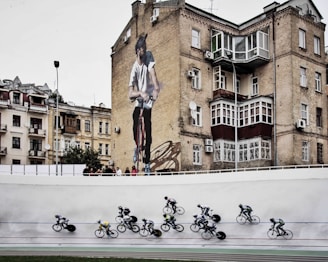 Cyclists are racing on a track in the foreground while an urban setting with older buildings serves as the backdrop, including a prominent mural of a man on a bicycle on the side of a brick building.