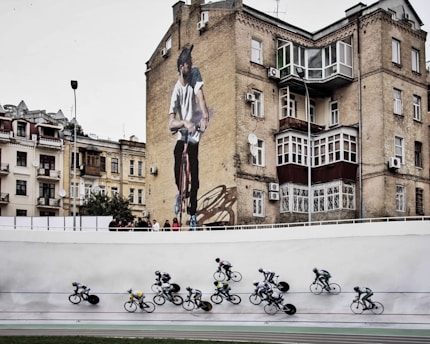 Cyclists are racing on a track in the foreground while an urban setting with older buildings serves as the backdrop, including a prominent mural of a man on a bicycle on the side of a brick building.