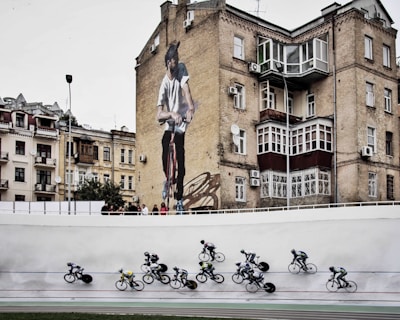Cyclists are racing on a track in the foreground while an urban setting with older buildings serves as the backdrop, including a prominent mural of a man on a bicycle on the side of a brick building.