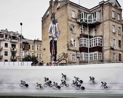 Cyclists are racing on a track in the foreground while an urban setting with older buildings serves as the backdrop, including a prominent mural of a man on a bicycle on the side of a brick building.