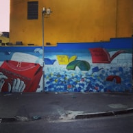 A colorful mural on a wall depicting a giant hand typing on a red typewriter. Papers scatter from the typewriter, transforming into books flying through the air. The titles of several authors and works, such as Machado de Assis and Charles Bukowski, are visible. The mural is set against a vivid yellow wall with a dark blue section and scattered multicolored balls below.