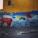 A colorful mural on a wall depicting a giant hand typing on a red typewriter. Papers scatter from the typewriter, transforming into books flying through the air. The titles of several authors and works, such as Machado de Assis and Charles Bukowski, are visible. The mural is set against a vivid yellow wall with a dark blue section and scattered multicolored balls below.