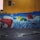 A colorful mural on a wall depicting a giant hand typing on a red typewriter. Papers scatter from the typewriter, transforming into books flying through the air. The titles of several authors and works, such as Machado de Assis and Charles Bukowski, are visible. The mural is set against a vivid yellow wall with a dark blue section and scattered multicolored balls below.
