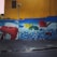 A colorful mural on a wall depicting a giant hand typing on a red typewriter. Papers scatter from the typewriter, transforming into books flying through the air. The titles of several authors and works, such as Machado de Assis and Charles Bukowski, are visible. The mural is set against a vivid yellow wall with a dark blue section and scattered multicolored balls below.