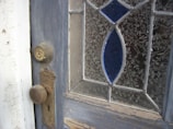 A close-up view of a weathered wooden door featuring a vintage doorknob and lock. The glass panel in the door is intricately patterned with floral designs and highlighted with blue stained glass inserts.