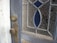 A close-up view of a weathered wooden door featuring a vintage doorknob and lock. The glass panel in the door is intricately patterned with floral designs and highlighted with blue stained glass inserts.