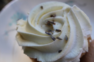 A close-up of a cupcake topped with creamy white frosting, swirled into a rose shape. Small pieces of dried lavender are sprinkled on top, adding a touch of natural texture.