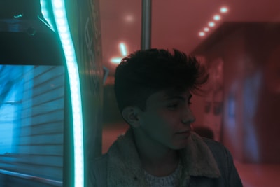 A person with short hair is viewed from the side, standing near a bright, vertical strip of neon light. The setting is indoors with a warm, ambient glow from ceiling lights reflecting on smooth surfaces. The person wears a coat with a textured interior lining.