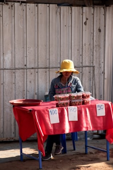 A person wearing a yellow hat and a plaid apron is sitting at a makeshift market stall. The table is covered with a red cloth and several containers of strawberries are displayed for sale, with price tags of 100, 80, and 50. The background consists of a corrugated metal wall.