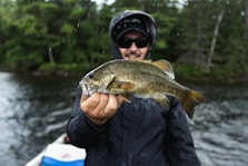 A person holding a freshly caught fish, standing on a boat. The individual is wearing a dark rain jacket and sunglasses, suggesting a cool or rainy day. The background consists of a body of water surrounded by dense green trees.