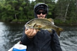 A person holding a freshly caught fish, standing on a boat. The individual is wearing a dark rain jacket and sunglasses, suggesting a cool or rainy day. The background consists of a body of water surrounded by dense green trees.