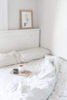 A cozy bedroom corner with a neatly made bed featuring soft white bedding. A wooden headboard holds a framed art piece, and a light wood tray resting on the bed contains a smartphone and a mug filled with frothy coffee.