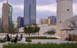 A cityscape featuring modern skyscrapers and a historical tower amidst a public space with greenery. People are casually sitting and walking, suggesting a mix of contemporary urban life and cultural heritage.