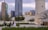A cityscape featuring modern skyscrapers and a historical tower amidst a public space with greenery. People are casually sitting and walking, suggesting a mix of contemporary urban life and cultural heritage.