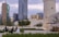 A cityscape featuring modern skyscrapers and a historical tower amidst a public space with greenery. People are casually sitting and walking, suggesting a mix of contemporary urban life and cultural heritage.