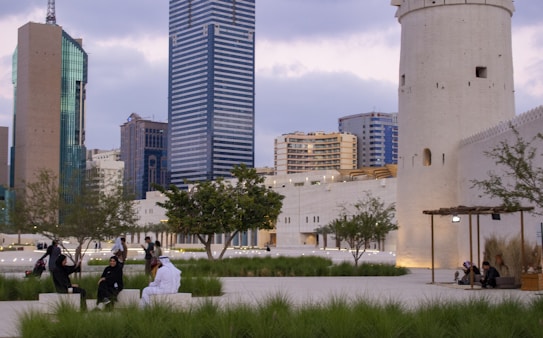 A cityscape featuring modern skyscrapers and a historical tower amidst a public space with greenery. People are casually sitting and walking, suggesting a mix of contemporary urban life and cultural heritage.