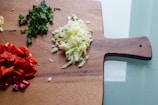 A wooden cutting board holds finely chopped vegetables. There are red bell peppers, white onions, and green leafy herbs neatly arranged on the board, indicating meal preparation.