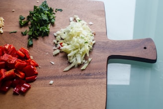A wooden cutting board holds finely chopped vegetables. There are red bell peppers, white onions, and green leafy herbs neatly arranged on the board, indicating meal preparation.
