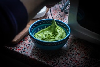 A blue bowl filled with a creamy green mixture is being stirred by a person using a spoon. The bowl is placed on a countertop with a red and black terrazzo pattern. A microwave is partially visible to the right, and a wooden cutting board is seen in the background.