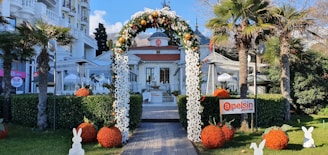 An elegant outdoor entrance with an arch decorated with flowers and ornaments. The path leads to a stylish cafe, surrounded by palm trees and trimmed hedges. Decorative orange balls and rabbit figures enhance the inviting garden setting.