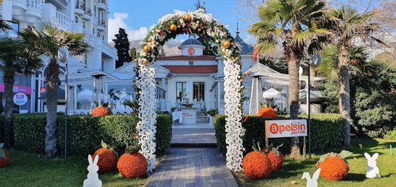 An elegant outdoor entrance with an arch decorated with flowers and ornaments. The path leads to a stylish cafe, surrounded by palm trees and trimmed hedges. Decorative orange balls and rabbit figures enhance the inviting garden setting.