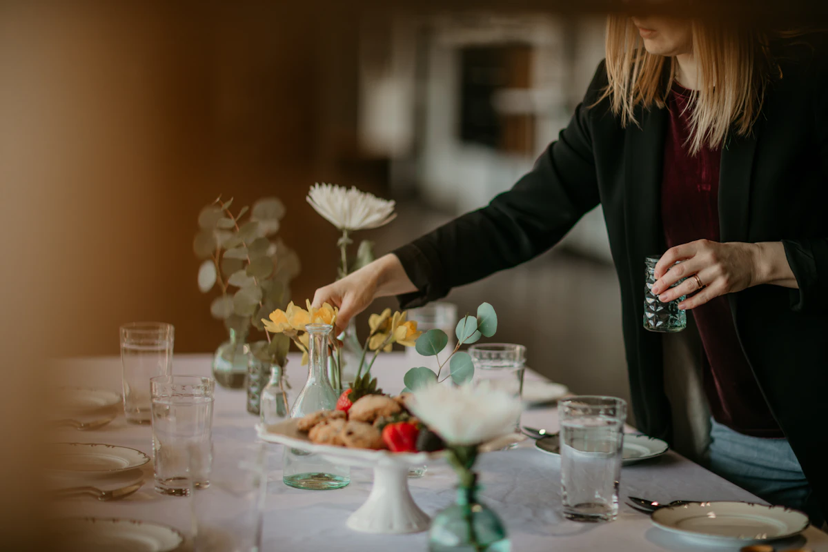 Une femme en veste sombre pose un petit vase de fleurs sur une table dressée, entre les verres, les assiettes et les compositions déjà installées