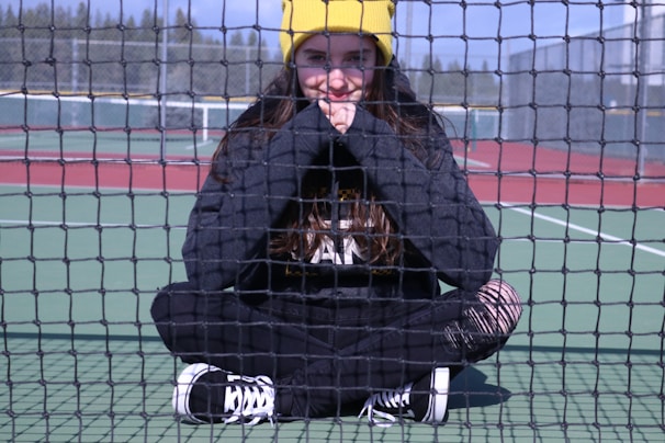 A person is sitting cross-legged on a tennis court, behind a net. They are wearing a black outfit with white sneakers and a bright yellow beanie. The background includes tennis court markings and a fence with trees visible beyond.