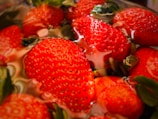 Fresh strawberries partially submerged in water, showcasing their vibrant red color and textured surface with green leaves still attached. The water creates a reflective, glossy effect on the strawberries.